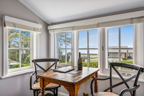 Una habitación con una mesa y sillas de madera, ventanas amplias con persianas, y una botella sobre la mesa.