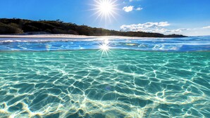 Ein Strand mit klarem türkisblauem Wasser, weißem Sand und einer bewaldeten Hügelkuppe im Hintergrund.