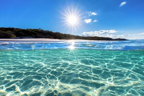 Una playa con aguas turquesas cristalinas, una orilla de arena blanca y una colina boscosa al fondo.