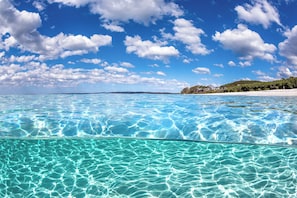 Mar azul y transparente con una playa arenosa y una colina boscosa al fondo.