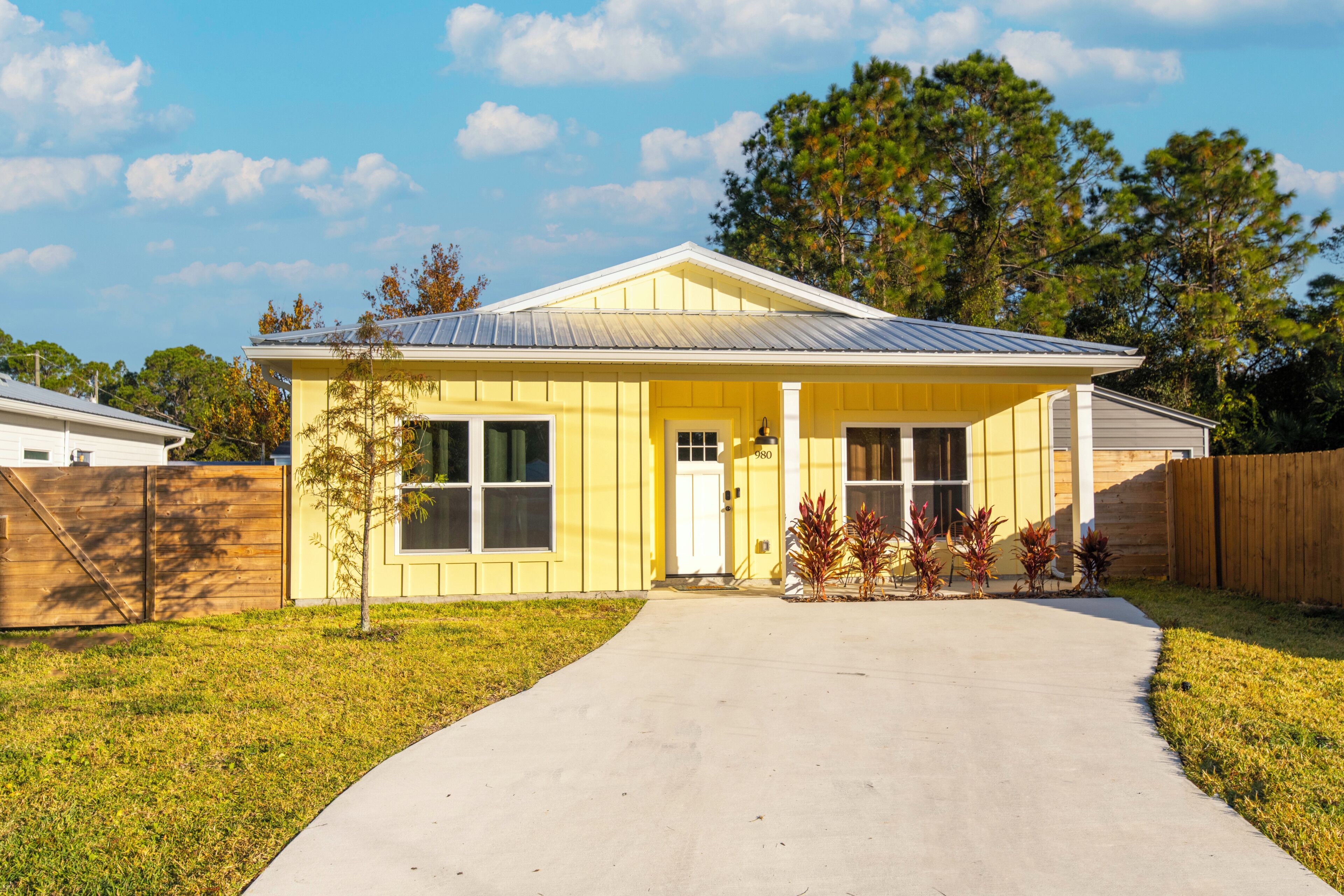 A single-story yellow house with a front porch, a small tree, and a paved driveway.
