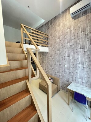 A staircase with wooden railings leading to a landing, a sofa, a table, and a wall with a herringbone pattern. - Apple 1 Condovilla (Jakarta Selatan)