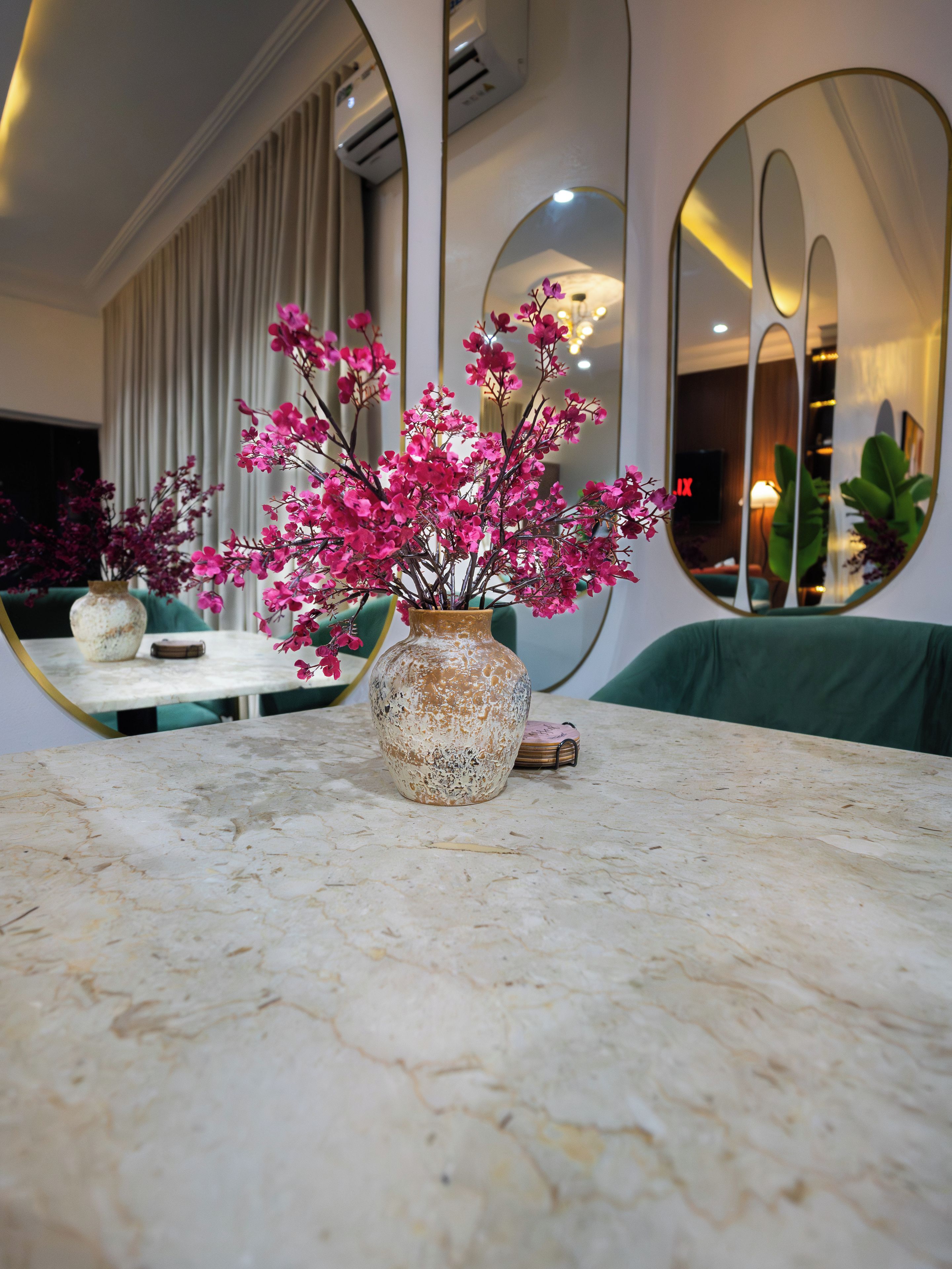 Une table en marbre surmontée d’un vase rempli de fleurs roses, un canapé vert et un grand miroir qui reflète la pièce.