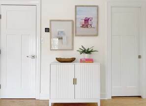 Eine weiße Tür mit Griff, ein weißes Schränkchen mit zwei Türen und goldenen Griffen, ein gerahmtes Bild an der Wand und eine kleine Pflanze auf dem Schränkchen.