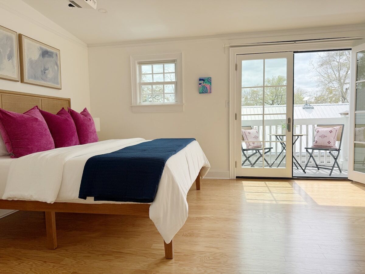 A bedroom with a bed, a balcony with chairs, and a view of the outside.