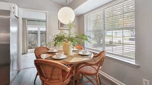 Een eethoek met een ronde tafel, rieten stoelen, een potplant en een raam met jaloezieën.