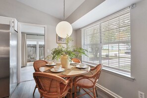 Un coin repas avec une table ronde, des chaises en osier, une plante en pot et une fenêtre avec des stores.