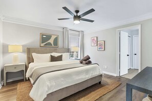 A bedroom with a bed, nightstands, a ceiling fan, artwork, and a view of a hallway. - Halfway to Tybee Beach and Downtown Savannah: Renovated Cottage, Dogs Welcome (Savannah)