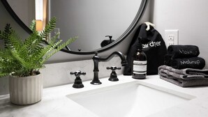 Een badkamer met een wit wastafel, zwarte kranen, een spiegel, een potplant, een handzeepfles en handdoeken met de tekst 'air dryer' en 'makeup'.