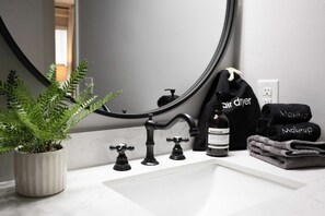 Une salle de bain avec un lavabo blanc, des robinets noirs, un miroir, une plante en pot, un flacon de savon liquide et des serviettes étiquetées « sèche-cheveux » et « maquillage ».