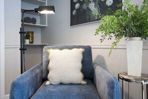 Een stoel met een decoratief kussen, een vloerlamp, een potplant en een kunstwerk in een lijst aan de muur.
