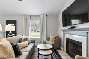 Een woonkamer met een bank, fauteuil, open haard, televisie en een raam met gordijnen.