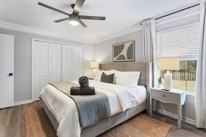 Une chambre à coucher comprenant un lit, des tables de chevet, un ventilateur de plafond, une fenêtre avec des stores et une porte.