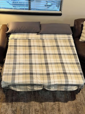 A bed with a checkered bedspread, two pillows, a window with blinds, and a brown leather chair. - Escape to the pines at Bow String Retreat (Munds Park)