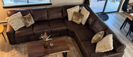 A brown leather sectional sofa with decorative pillows, a wooden coffee table with a vase, and a rustic rug.