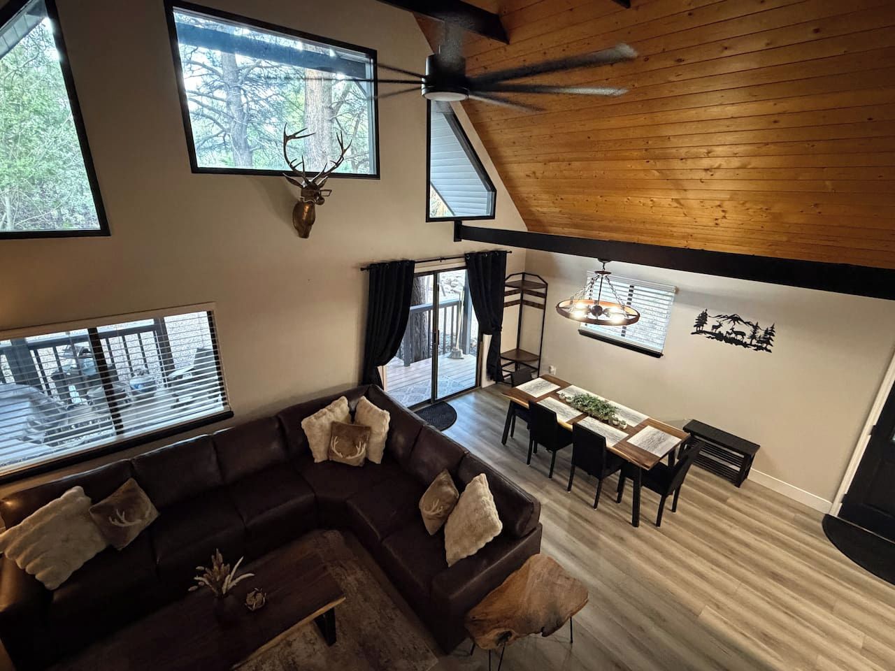A modern living room with a sectional sofa, dining table, and chairs, large windows with blinds, and a ceiling fan.