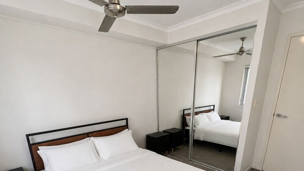 A neatly made bed with white linens and a rolled towel, a ceiling fan, and a large mirror reflecting the room. - River Glimpse 2BR Walk to CBD & Parking (Brisbane City)