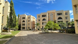 A paved courtyard with buildings on either side, a large tree, and a clear blue sky. - Pristine & Breezy 2BR Retreat with Pool, Gym & Balcony Views – Mtwapa Coast (Mtwapa)