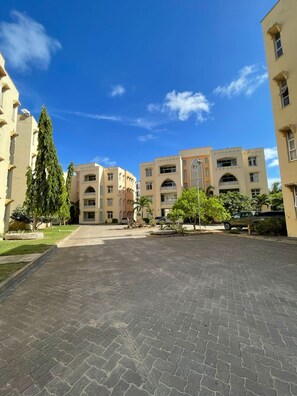 Um pátio pavimentado com edifícios em ambos os lados, uma grande árvore e um céu azul claro.