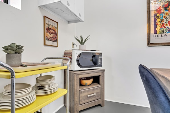 A kitchen area with a microwave on a wooden cabinet, a yellow shelf with plates, a potted plant, and a framed picture on the wall.