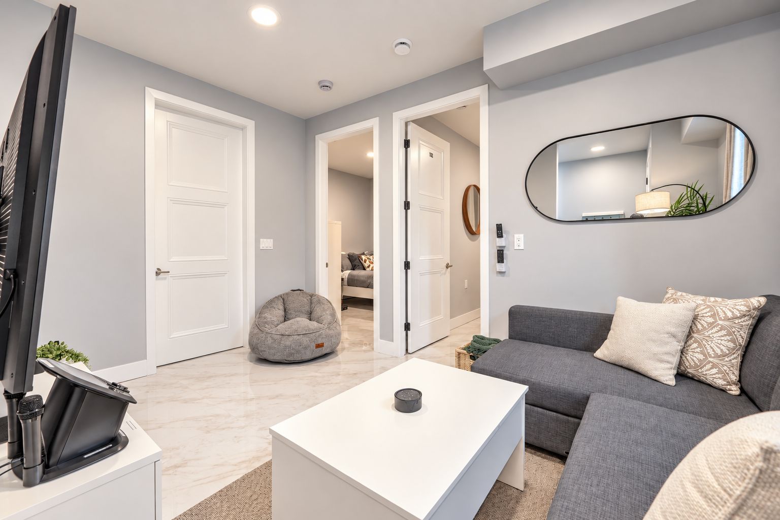 Un salon moderne avec un canapé gris, une table basse blanche, un fauteuil en forme de poire et un téléviseur à écran plat posé sur un meuble.