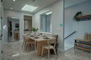 A modern dining area with a rectangular table set for four, flanked by two chairs. A large window with a view of the outside is visible, and a plant is placed on a shelf above the table.