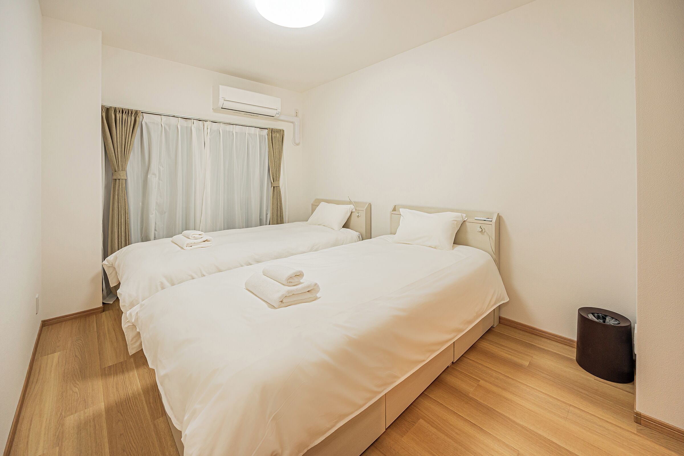 A hotel room with two beds, a waste bin, and an air conditioning unit.