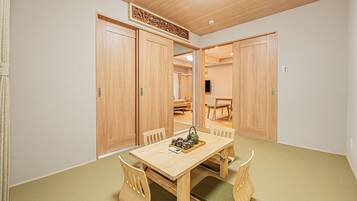 A room with a wooden dining table, chairs, and a view into another room with a television.