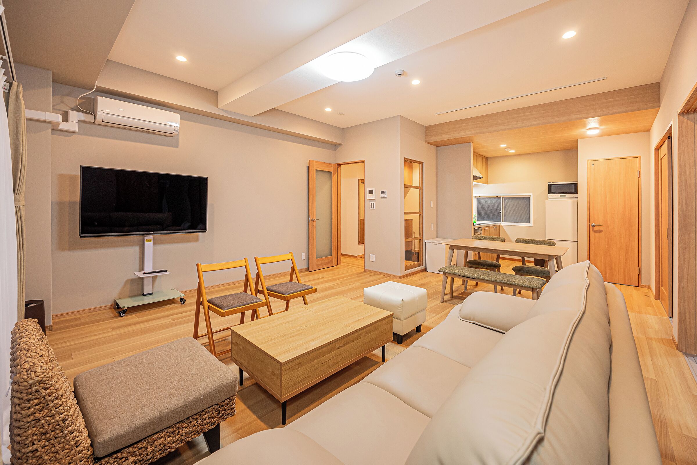 A modern living room with a flat-screen TV, wooden furniture, and a kitchen area in the background.