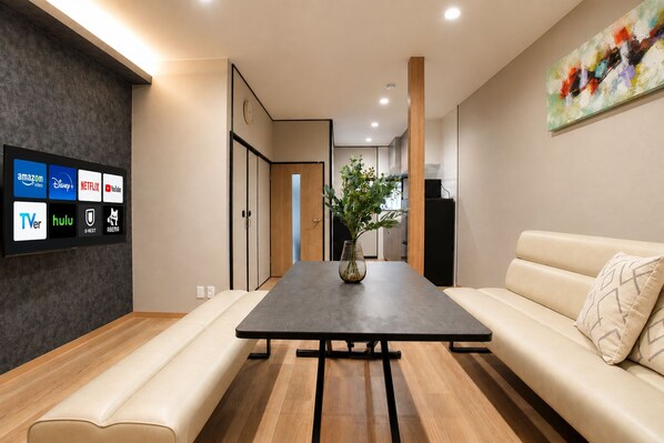 A modern living room with a flat-screen TV displaying streaming services, a wooden coffee table, a beige sofa, and a plant. - Nishikujo -EN- (Osaka konohanaku nishikujyo)