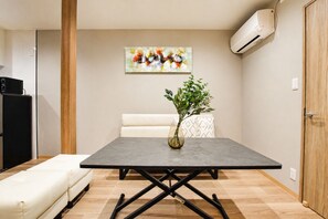 A modern living room with a grey table, a white sofa, a vase with green plants, and a colorful abstract painting on the wall. - Nishikujo -EN- (Osaka konohanaku nishikujyo)