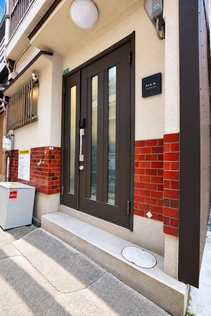A building entrance with a dark-colored door, a small step leading up to the entrance, and a red-tiled wall. - Nishikujo -EN- (Osaka konohanaku nishikujyo)