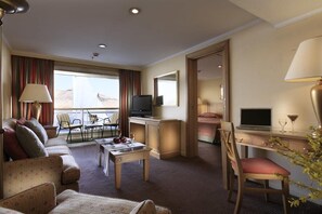 Et hotelværelse med sofa, spisebord, fjernsyn, skrivebord med en bærbar computer og udsigt til det fri.