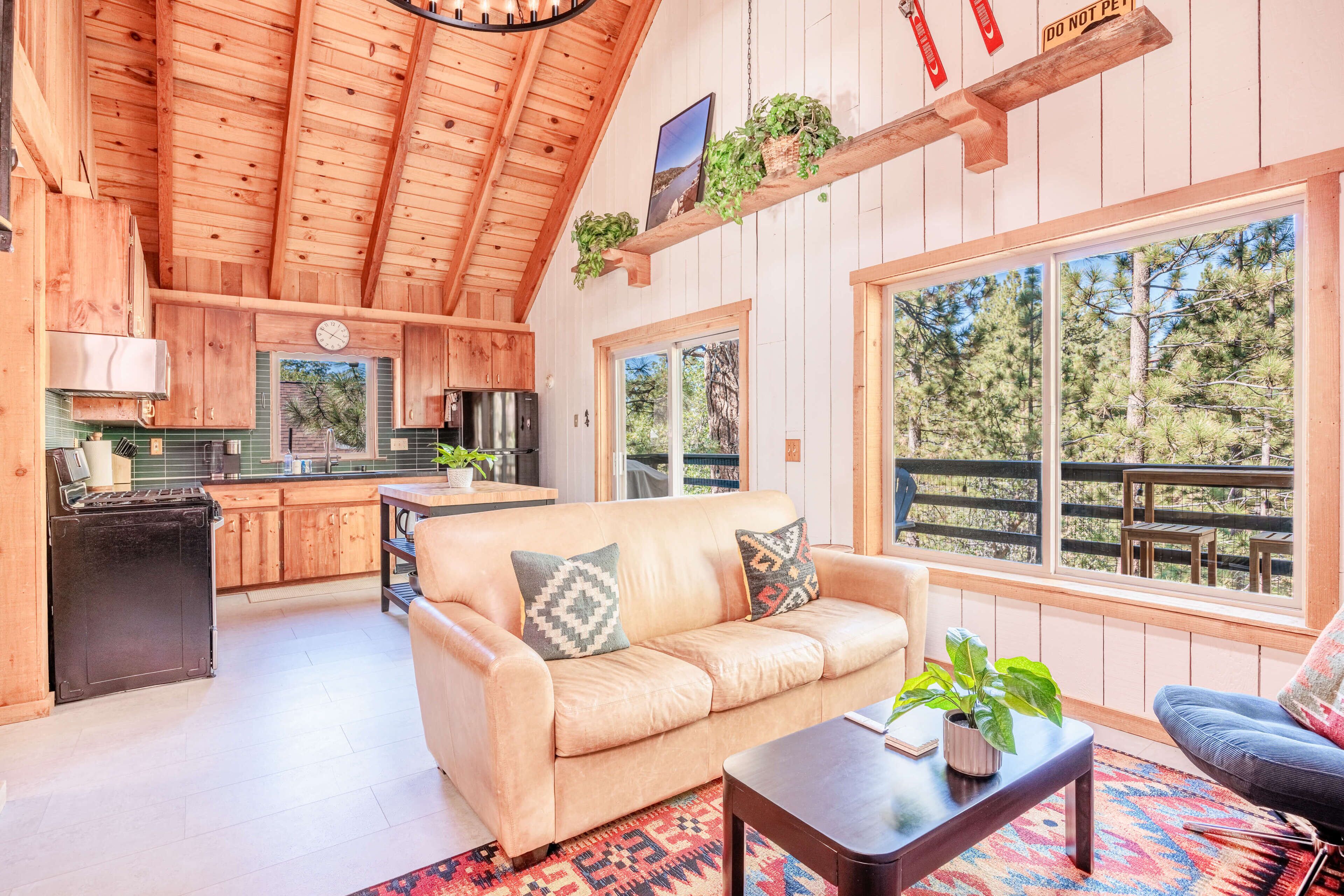 A cozy cabin interior with a wooden ceiling, a kitchen area, a living room with a sofa and coffee table, and large windows offering a view of trees.