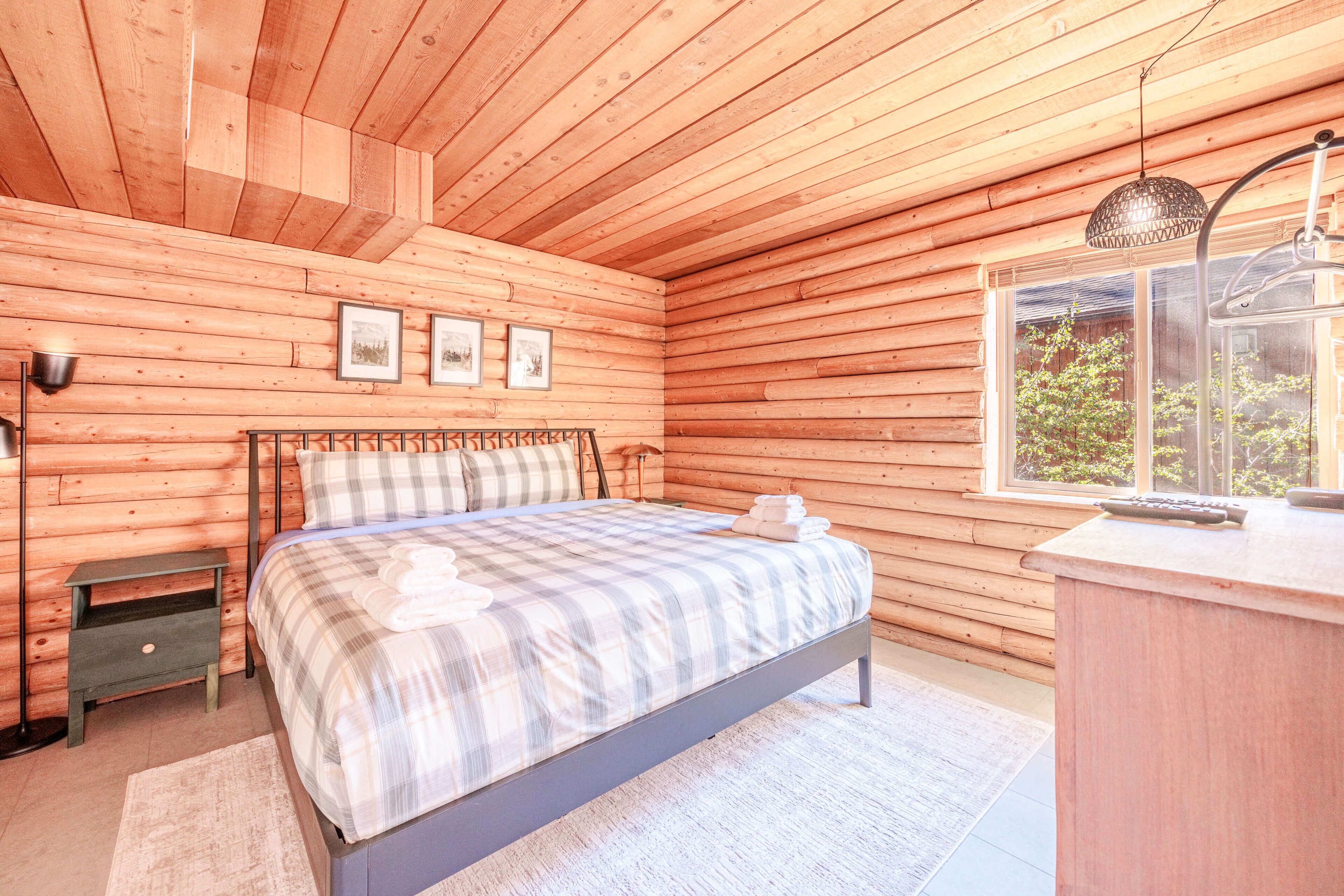 Un dormitorio de cabaña de madera con una cama, una mesita de noche, una lámpara y un ventanal.