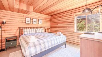 Un dormitorio de cabaña de madera con una cama, una mesita de noche, una lámpara y un ventanal.