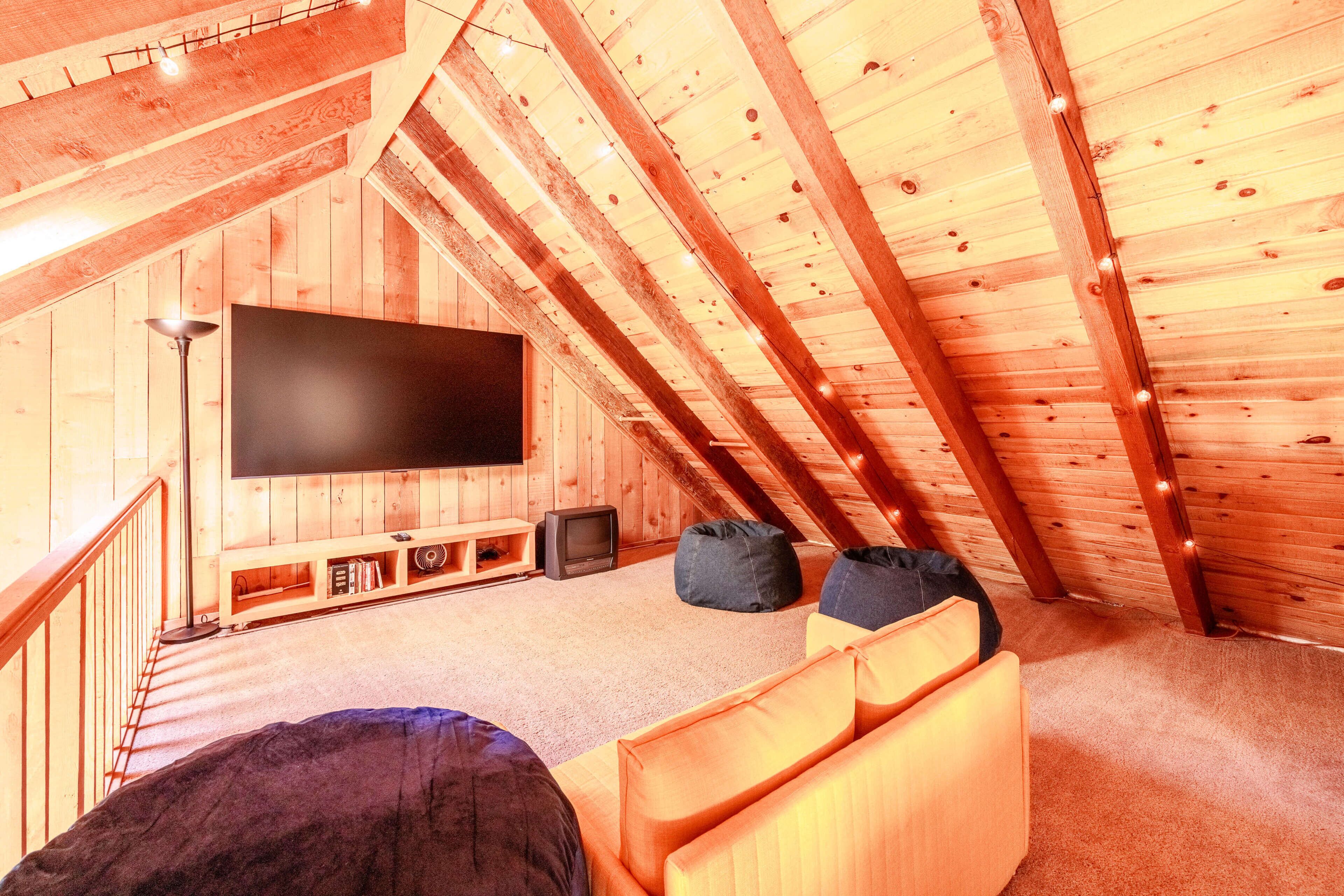 Habitación en el ático con techo de madera, un televisor de pantalla plana en la pared, un sofá y dos pufs.