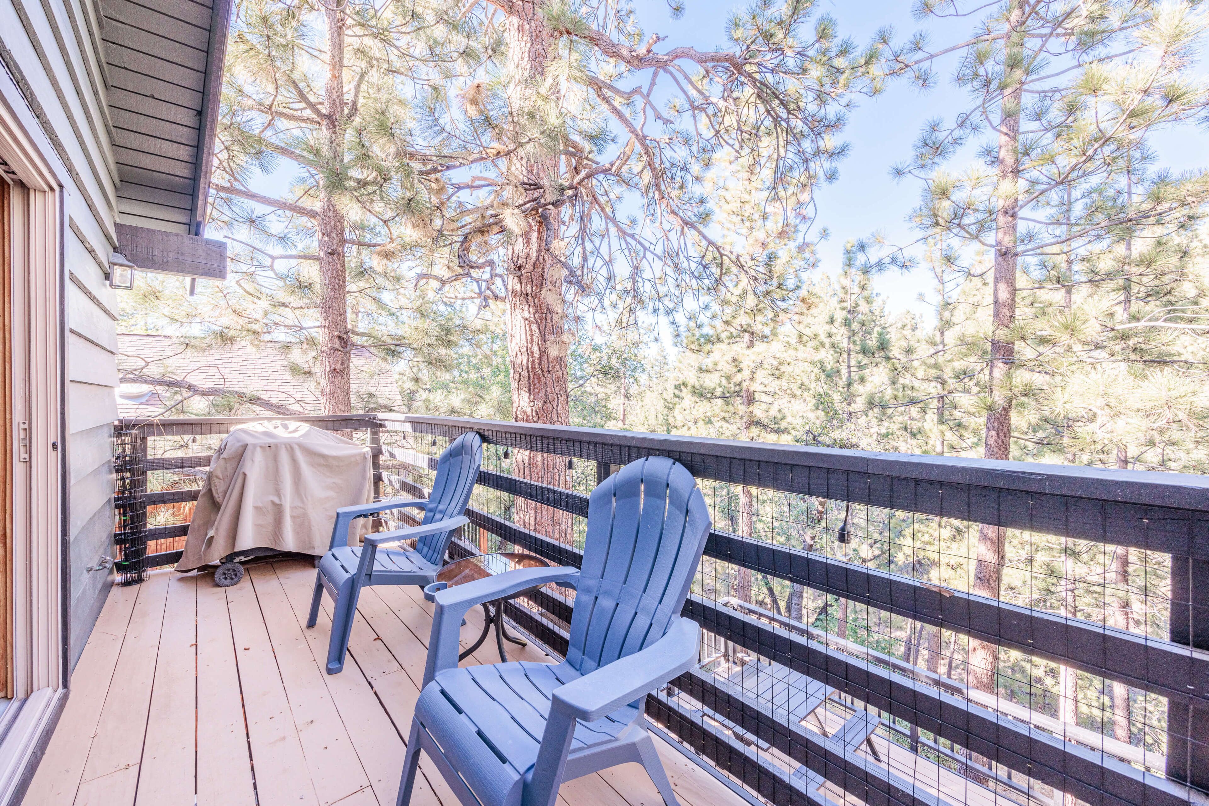 Une terrasse en bois avec deux fauteuils Adirondack, un barbecue abrité et une vue sur des pins.