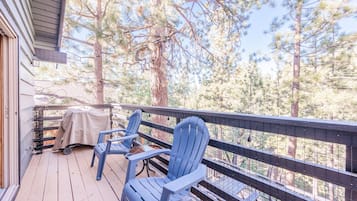Une terrasse en bois avec deux fauteuils Adirondack, un barbecue abrité et une vue sur des pins.