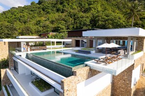 Una casa moderna con una gran piscina, mobiliario de exterior y vistas a una colina boscosa.