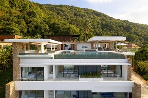 Una casa moderna de varios pisos con piscina en la azotea, barandillas de cristal y un exuberante paisaje de colinas verdes al fondo.