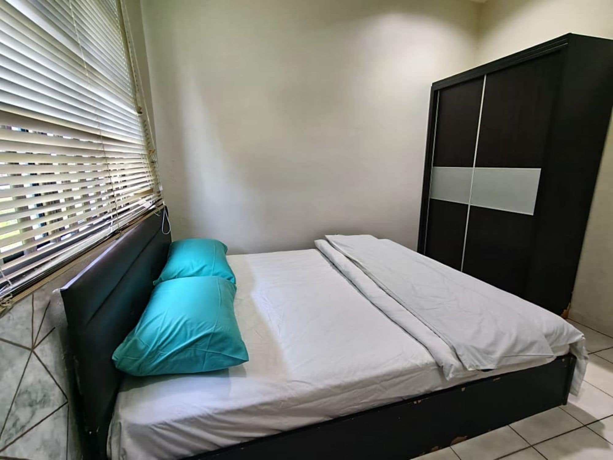 Uma cama de solteiro com colchão branco e almofada verde-água em quarto com piso ladrilhado e janela com persianas.