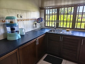 Cozinha com dispensador de água, chaleira, escorredor de louça e janela com vista para o verde.