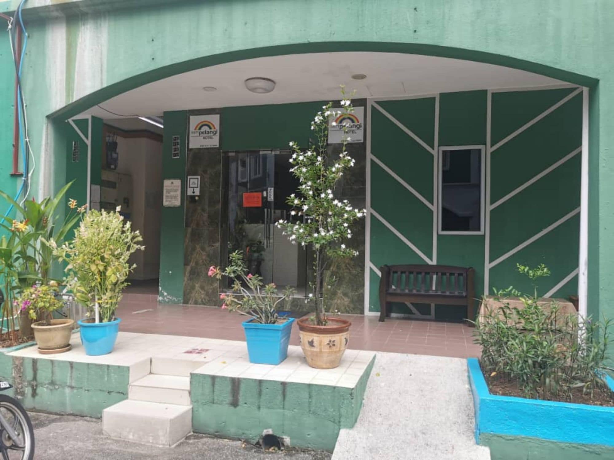 Entrada de hotel com fachada verde, plantas em vasos e um banco.