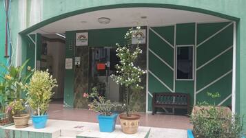 Entrada de hotel com fachada verde, plantas em vasos e um banco.