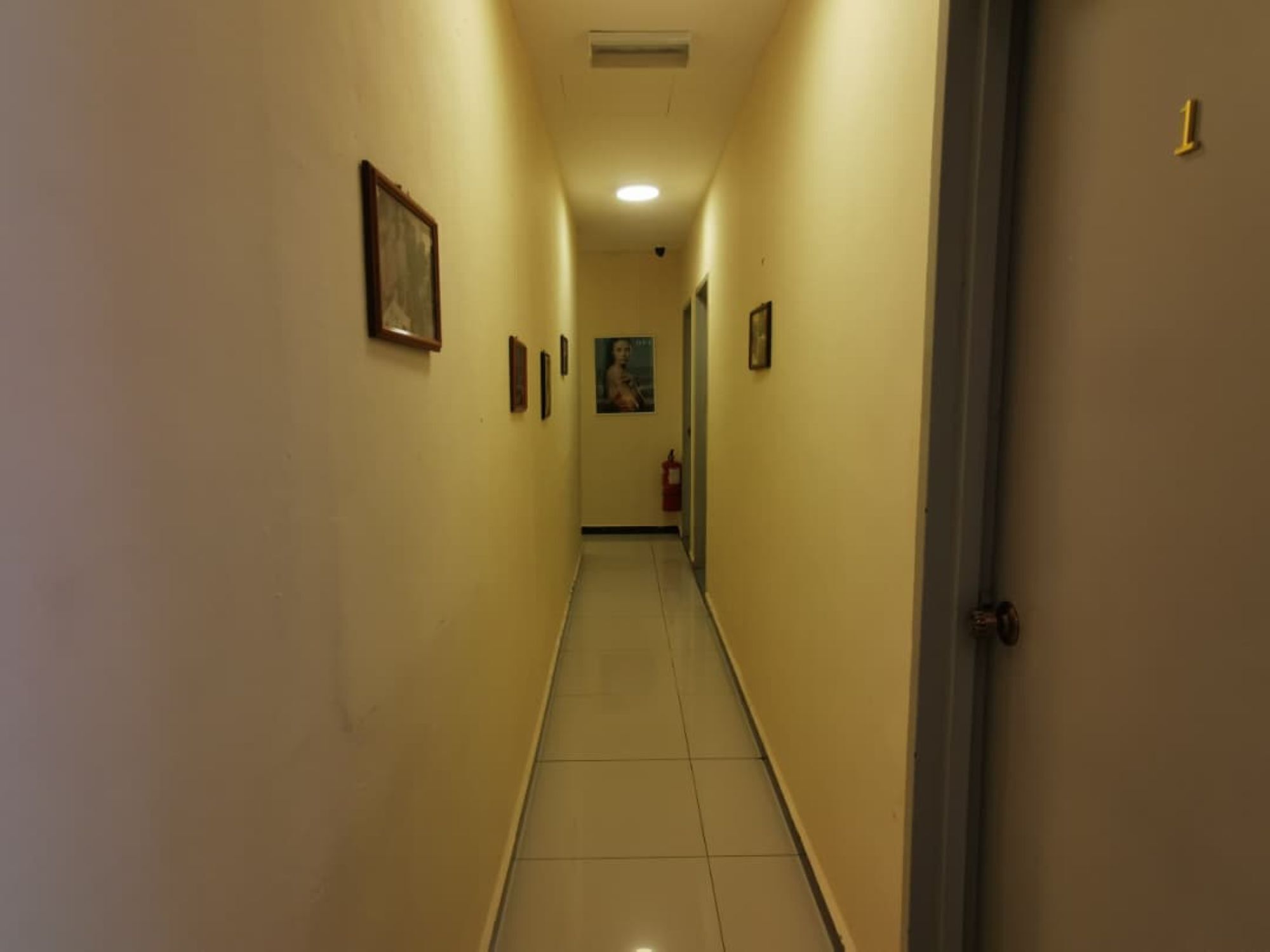 Um corredor estreito com piso de azulejos, uma porta à direita e várias imagens emolduradas nas paredes.