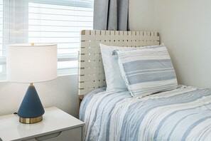 A neatly made bed with striped bedding, a bedside lamp, and a window with blinds. - Coconut Cottage | Beach Retreat Steps to Sand, Mini Golf & More (Indian Shores)