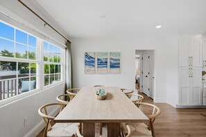 A dining area with a wooden table and chairs, a view of greenery outside, and a kitchen in the background. - Coconut Cottage | Beach Retreat Steps to Sand, Mini Golf & More (Indian Shores)