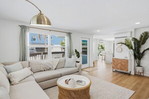 A modern living room with a sofa, a round coffee table, and a large window with blinds. - Coconut Cottage | Beach Retreat Steps to Sand, Mini Golf & More (Indian Shores)