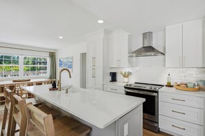 Una cocina moderna con armarios blancos, una isla central, una campana extractora de acero inoxidable y una zona de comedor con sillas de madera.